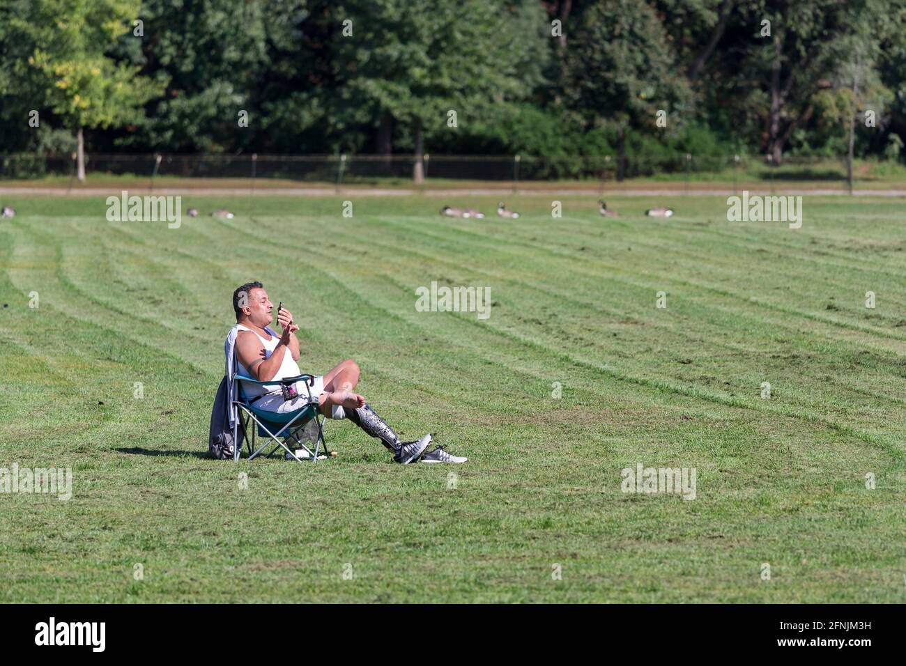 Man with prosthetic leg sitting in the chair in the field in summer, talking on mobile phone in NYC Stock Photo