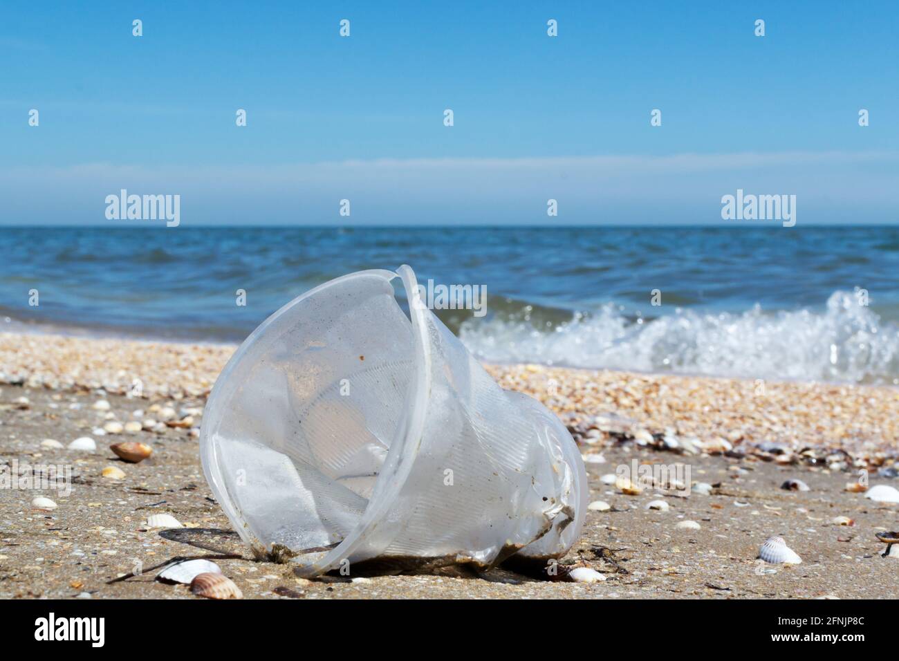 An old plastic cup lies on the seashore among seashells on the sand. Environmental pollution concept. Dirty sea. Ecological problem. Copy space. Stock Photo