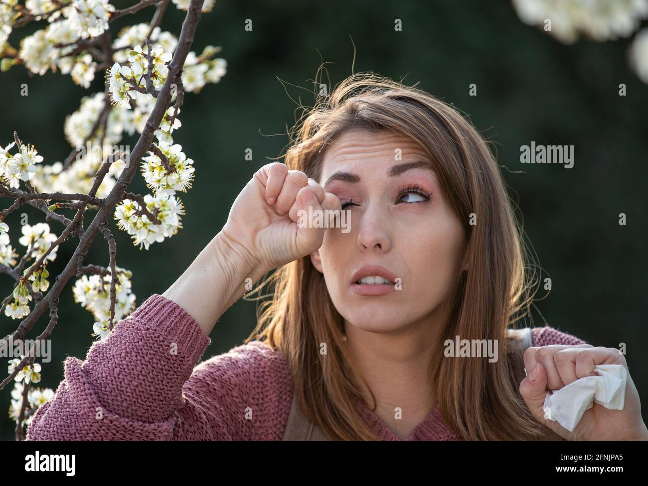Young pretty girl rubbing eyes beside blooming tree in spring time. Itchy eyes from allergy Stock Photo