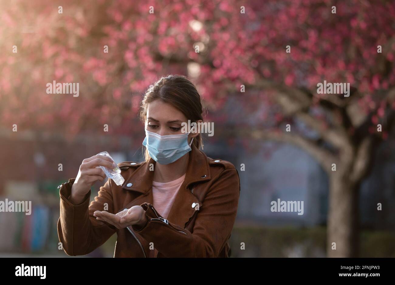 Young pretty woman with facial mask holding disinfectant gel and washing hands in front of blooming tree in spring Stock Photo