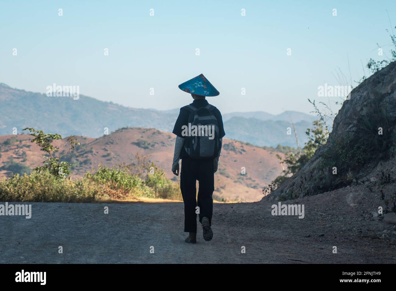 A local man in a blue hat and black traditional clothing walk a gravel road on a hike from Kalaw to Inle Lake, Shan state, Myanmar Stock Photo