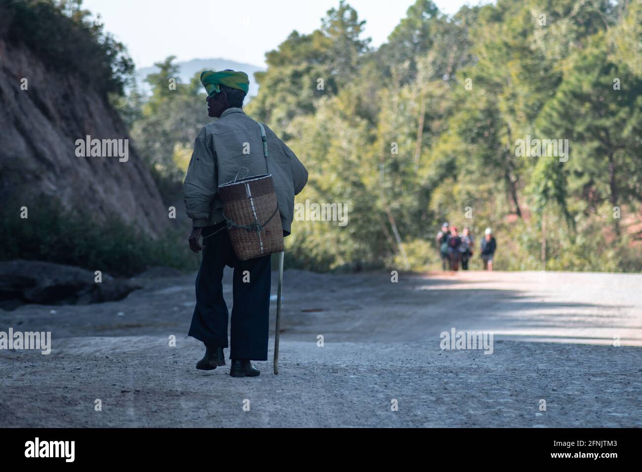 A local burmese man carries a basket on his back and walks down a gravel road with a walking stick between Kalaw and Inle Lake, Shan state, Myanmar Stock Photo