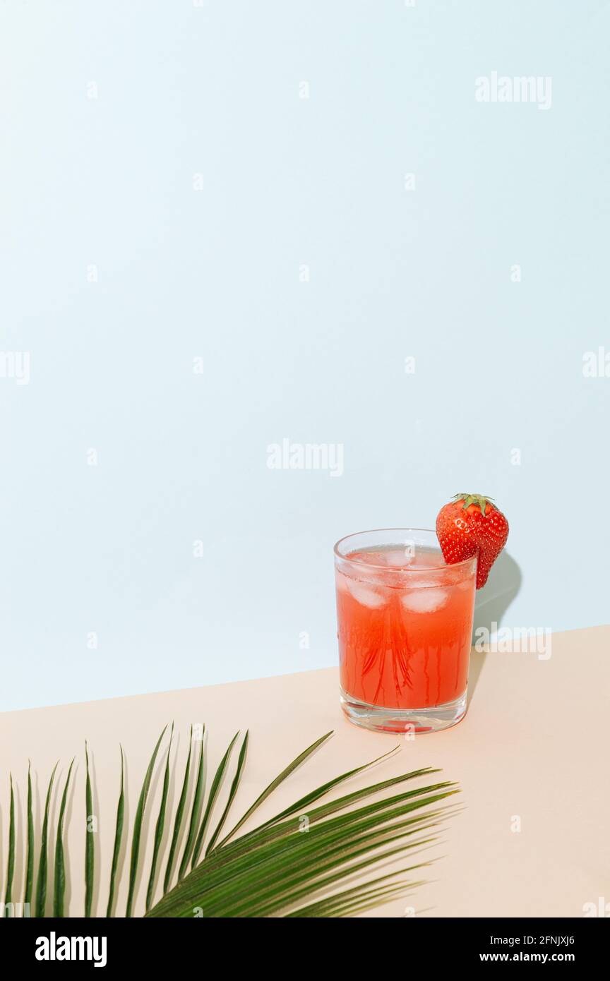 A fruity strawberry refreshing drink with ice. Tropical palm leaf blurred in front of the glass. Party summer refreshing concept on a combination of b Stock Photo