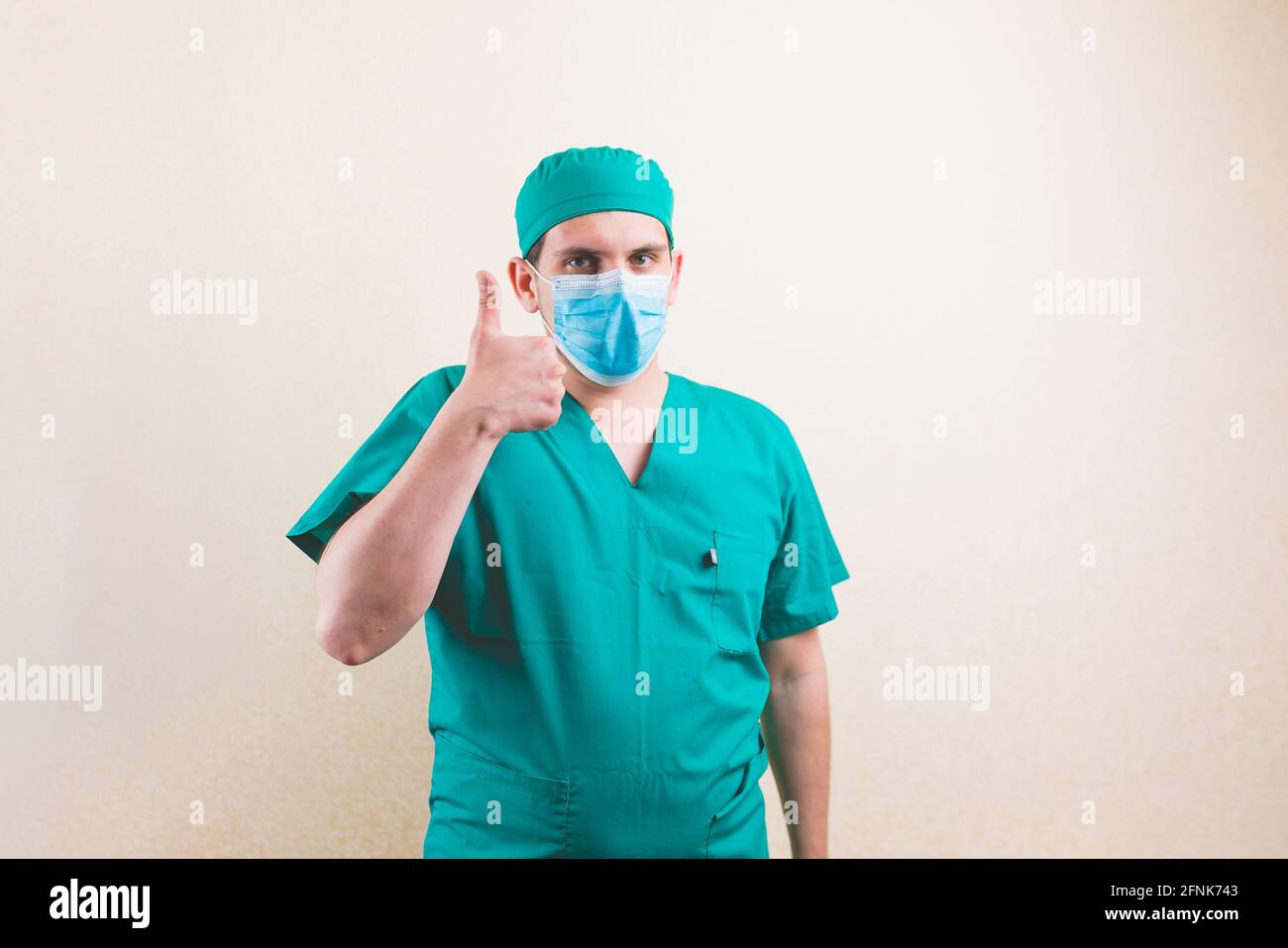 Young squinting doctor portrait with thumb up in green surgeon suit and protective breathing mask on the yellow background isolated Stock Photo