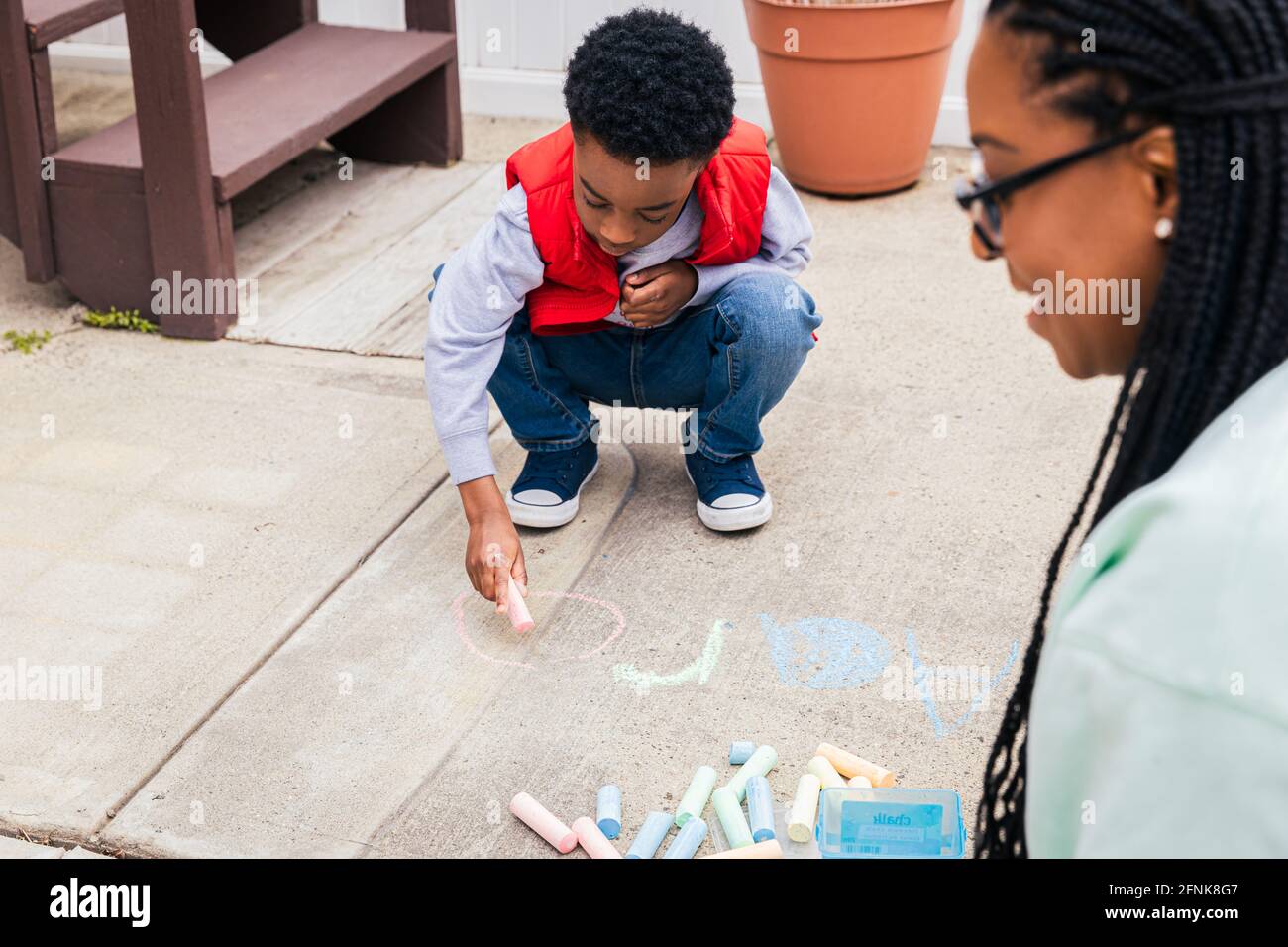 Boy using chalk to draw on concrete while mother smiles and helps Stock Photo