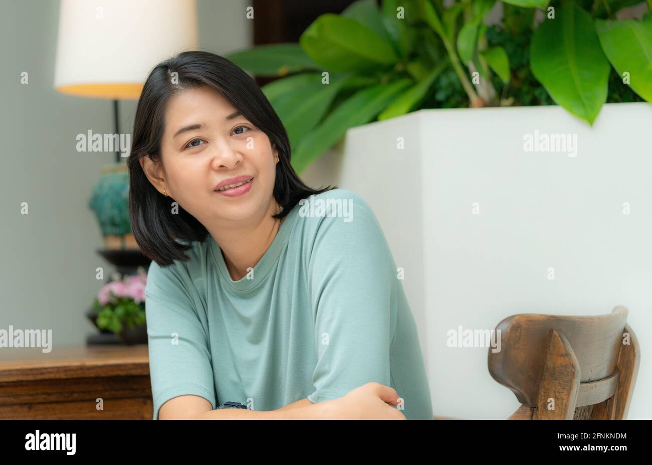 Portrait of beautiful Asian middle aged woman. Sitting on wooden chair in hotel's lobby, looking at camera. Stock Photo