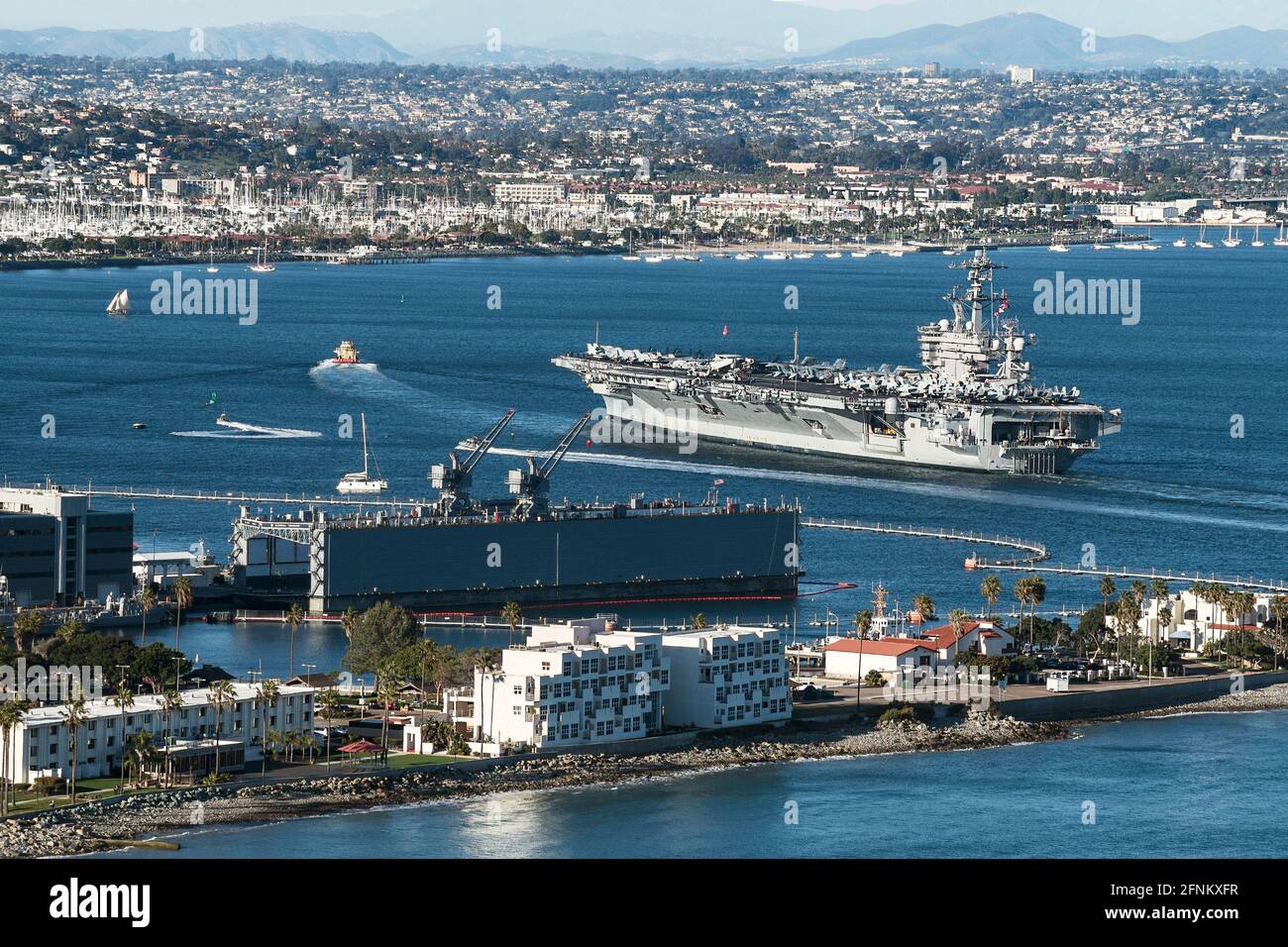 an aircraft carrier being led by a tugboat into San Diego Bay in California with a naval base in the foreground and the city of San Diego Stock Photo