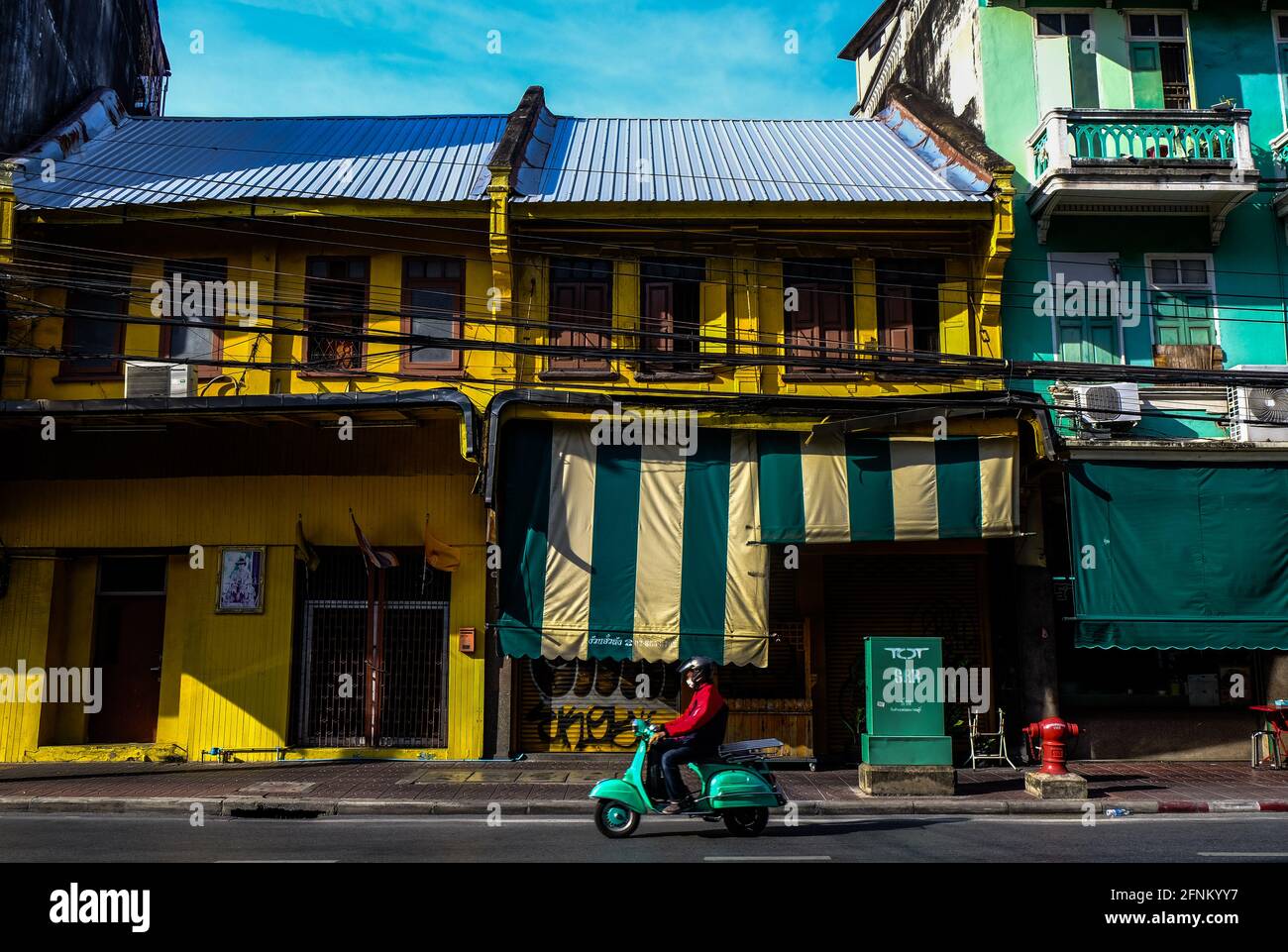 A man on an old green Vespa scooter rides past a restored yellow colored shopfront in the Chinatown area of Bangkok, Thailand Stock Photo