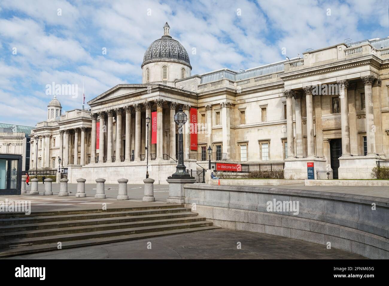 Two people outside the National Gallery taken from Trafalgar Square. One person sitting on a wall wearing a face mask, the other walking away. Stock Photo