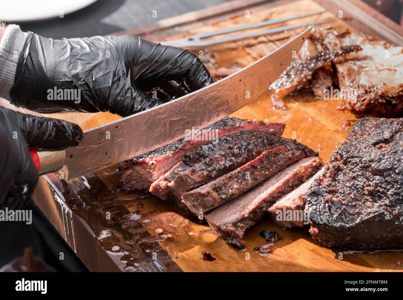 Gloved hands of a chef slicing a portion of delicious juicy tender roast beef brisket with a large knife in a close up on the blade and cutting board Stock Photo