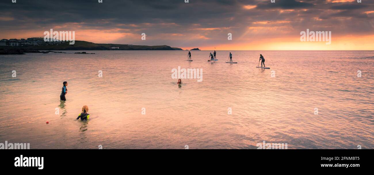 A panoramic image of children playing in the sea and a group of Stand Up Paddleboarders paddling across Fistral Bay in evening light at Newquay in Cor Stock Photo