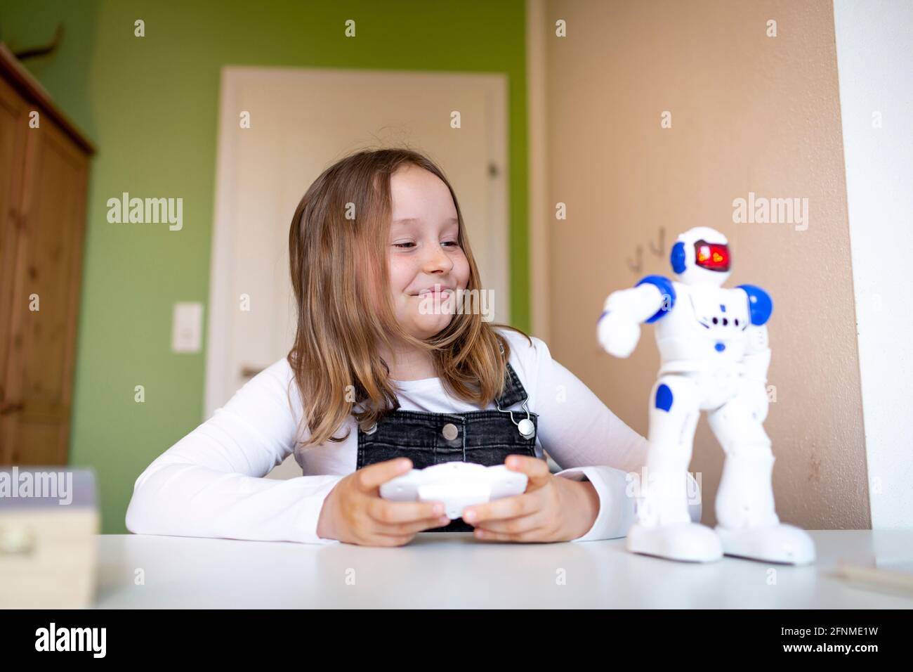 pretty young schoolgirl sitting on her desk in her room at home playing with small robot during corona time Stock Photo