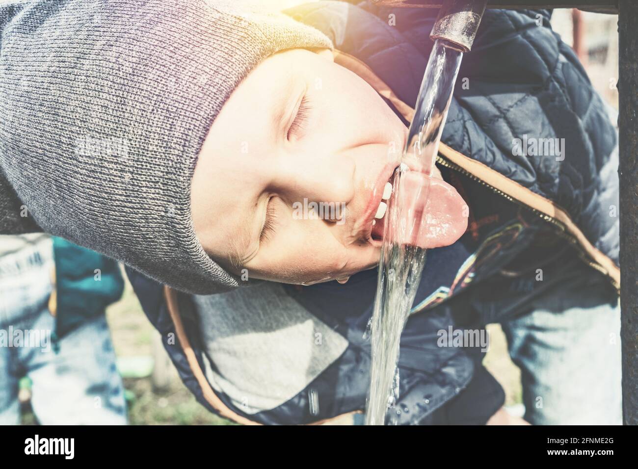 Funny little boy in grey knitted hat drinks water from vintage water pump in village house yard on sunny day close view Stock Photo