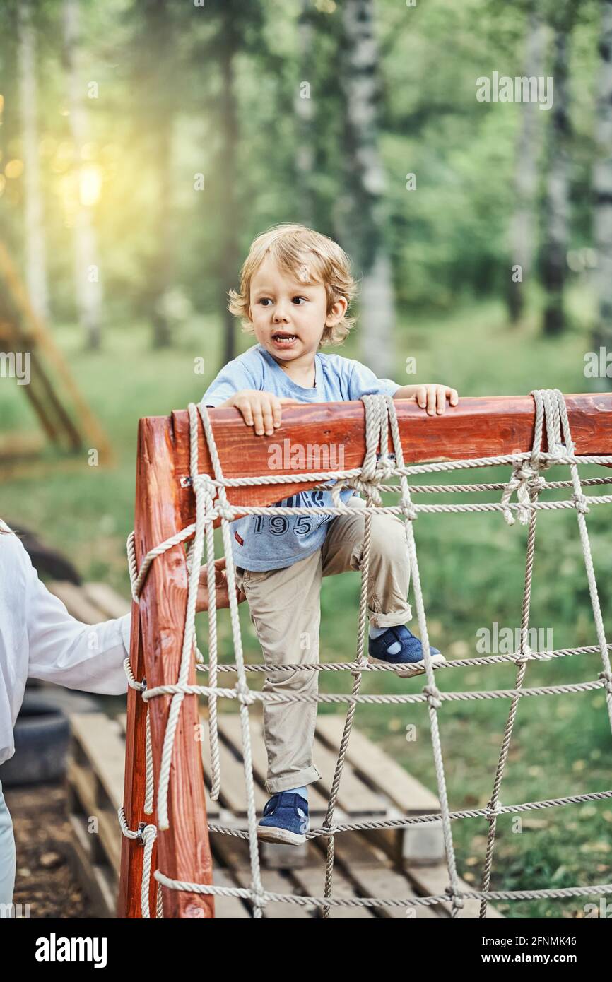 Playful little boy kid climbs onto obstacle with ropes under mother control on sports ground in green sunny summer city park Stock Photo