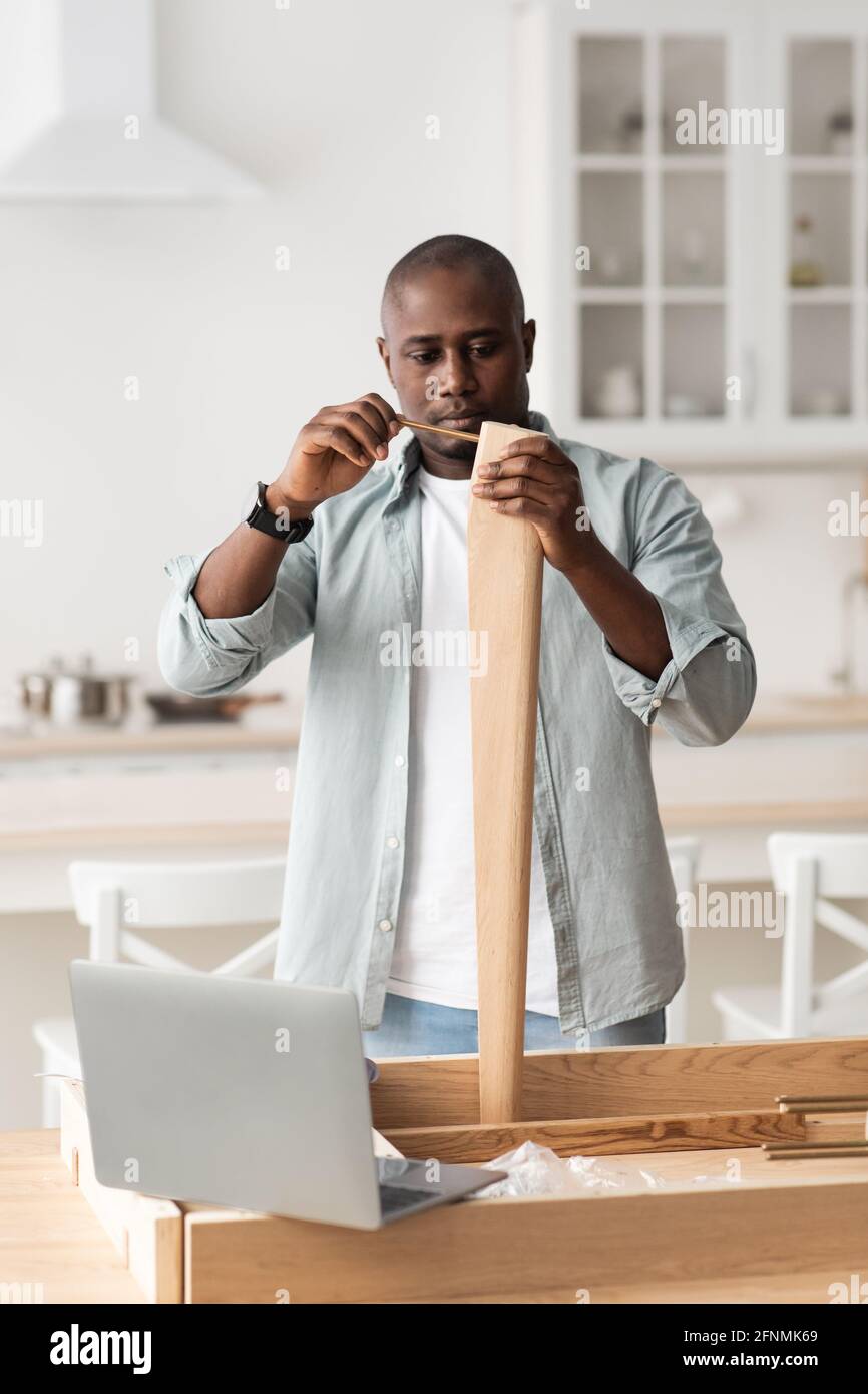 Modern handyman and online instructions. Black man screwing detail with manual screwdriver to table leg in kitchen Stock Photo