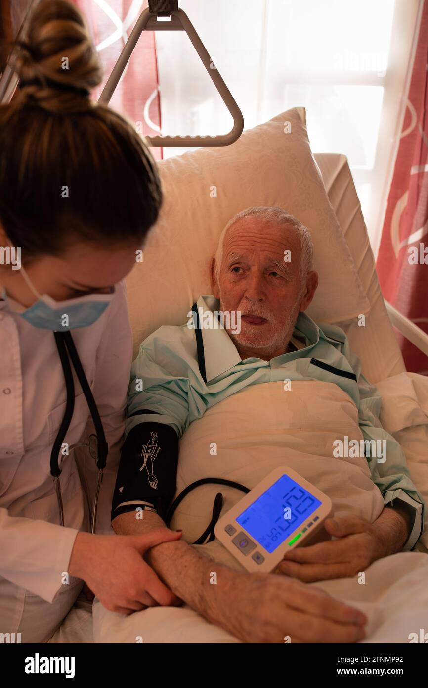 Young nurse measuring blood pressure to senior man lying in bed in hospital Stock Photo