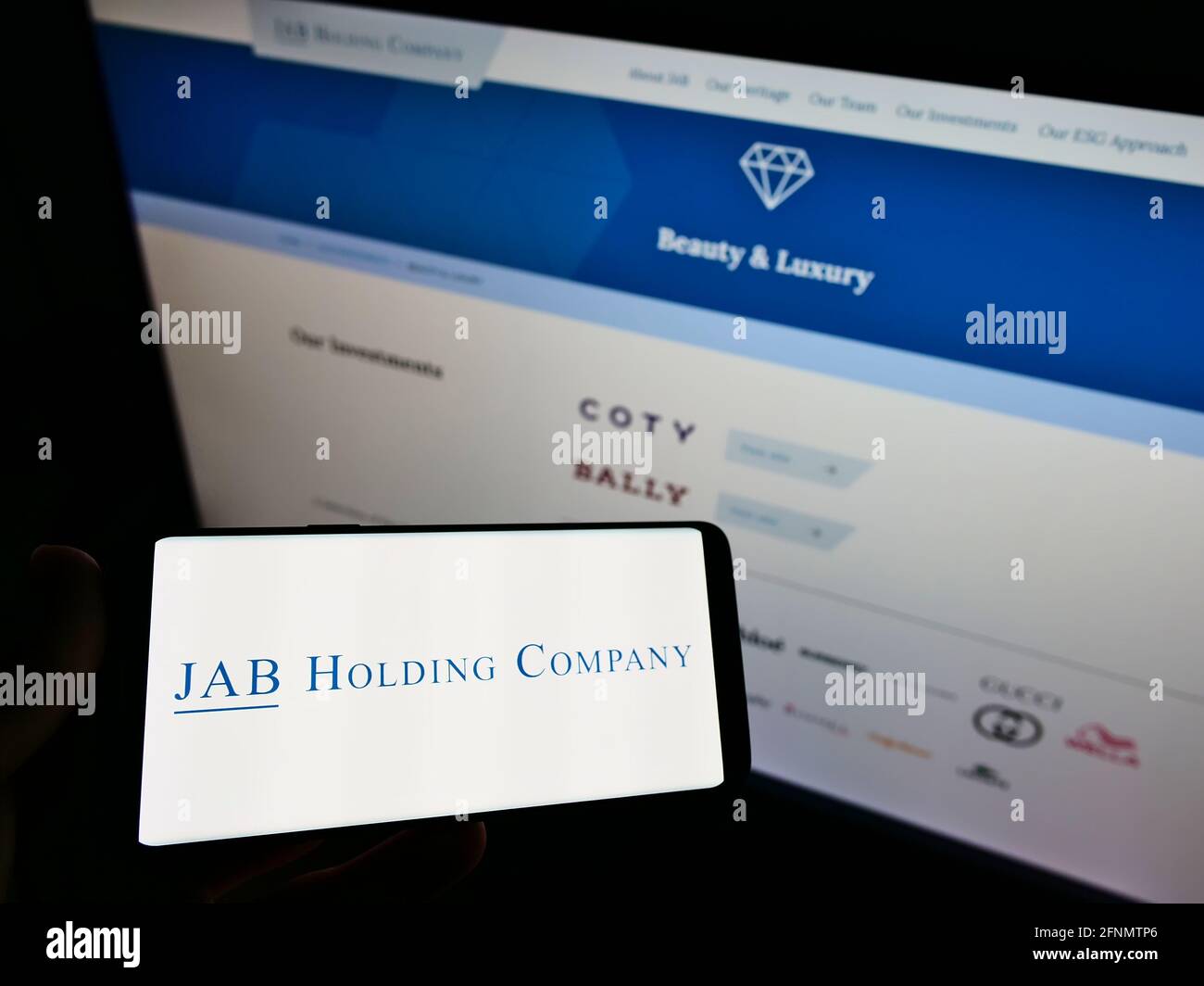 Person holding mobile phone with logo of business conglomerate JAB Holding Company S.à r.l. on screen in front of web page. Focus on phone display. Stock Photo