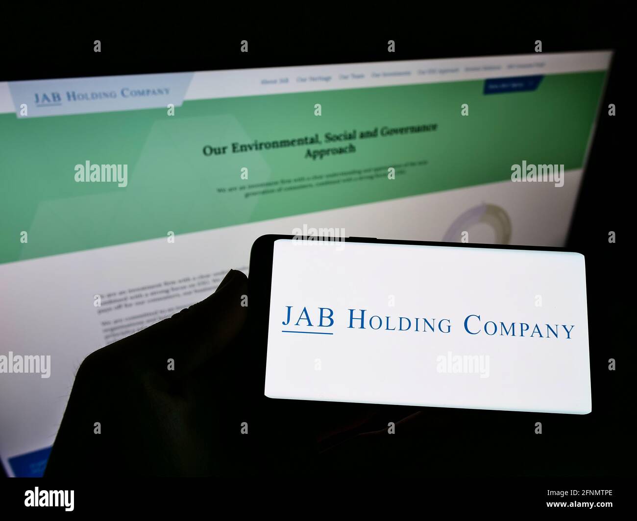 Person holding smartphone with logo of business conglomerate JAB Holding Company S.à r.l. on screen in front of webpage. Focus on cellphone display. Stock Photo