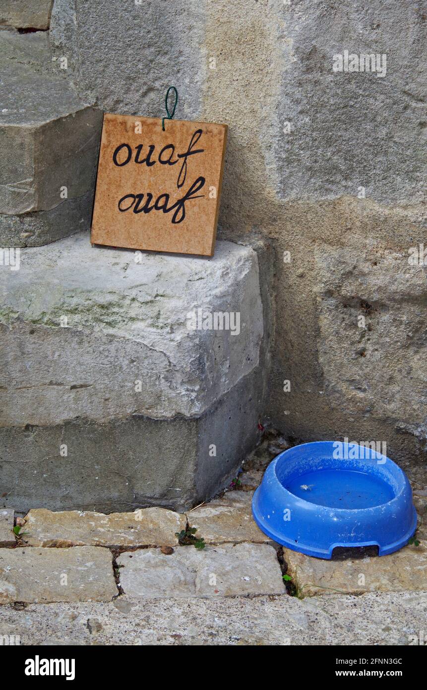 A small blue plastic dog’s drinking bowl and a sign saying “woof  woof” in french on the steep pedestrianised rue Montebello, Montmorillon, France Stock Photo