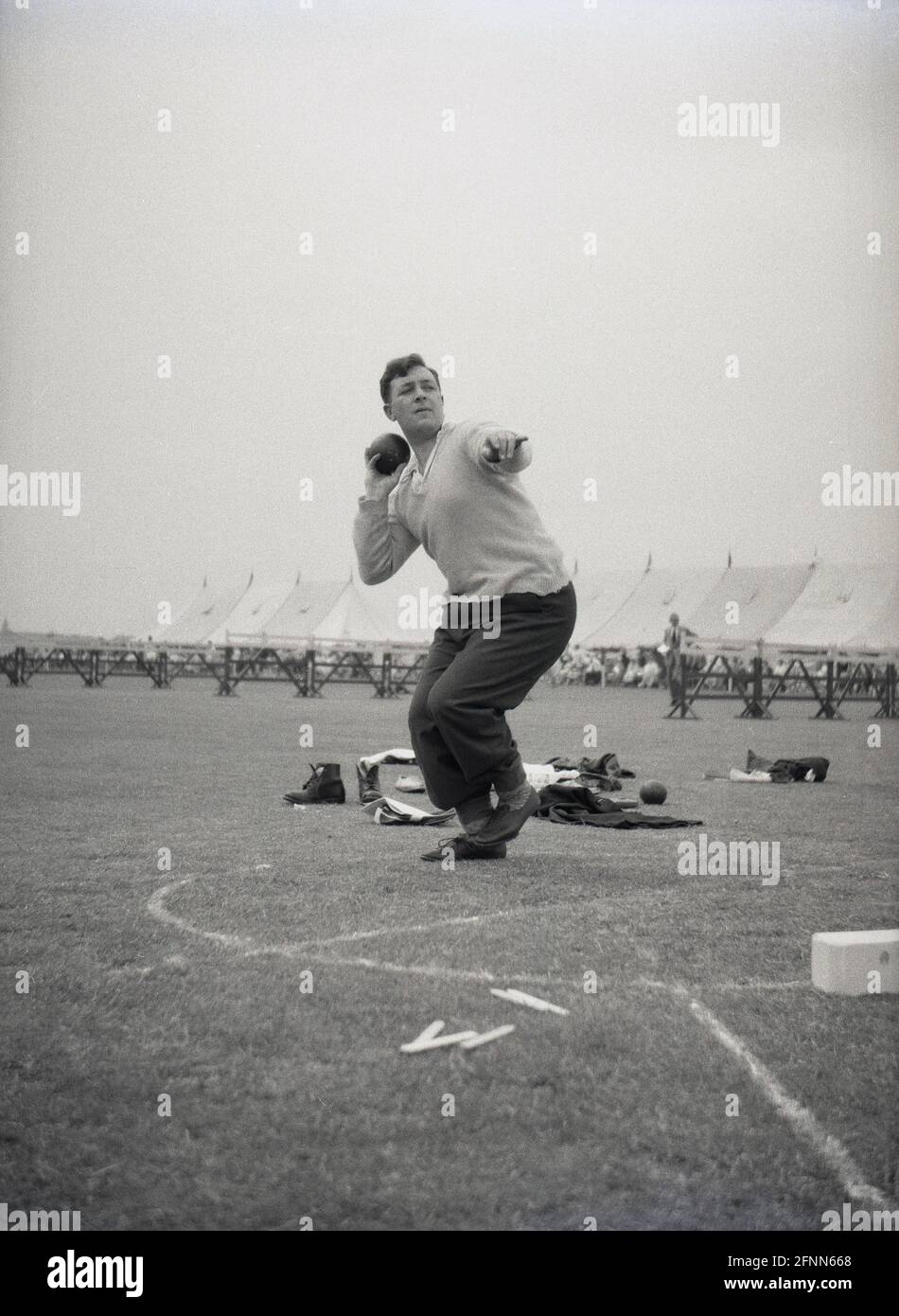 1954, historical, a man wearing a pair of trousers and a shirt and sweater, throwing the shotput, at a civil service sports day, England, UK, an amateur sports event. Putting the shot - a heavy metal ball - is a track and field event and has been part of the modern Olympic games since their revival in 1896. The shot put derives from the ancient sport of putting the stone. Stock Photo