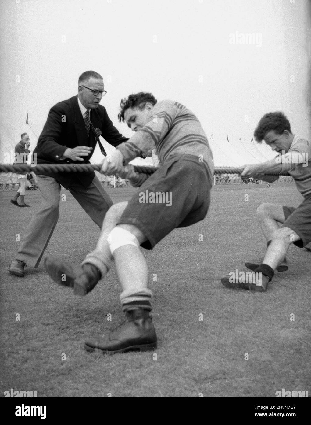 1954, historical, outside in a field, at a civil service sports day, in a tug-of-war contest, an official beside two male competitors wearing heavy ex-army boots pulling on the rope, checking whether they have crossed the winning point. An ancient sport, Tug of War which was in the summer Olympic games from 1900 to 1920, is a test of strength between two teams with the goal being to bring the rope a certain distance in one direction against the force of the opposite team's pull. Strong leg and arm muscles are needed. Stock Photo