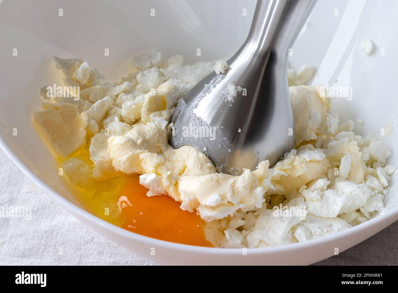 Cottage cheese, butter, egg and sugar in a white bowl - ingredients for mixing with a hand blender. The process of making curd bagels or croissants Stock Photo