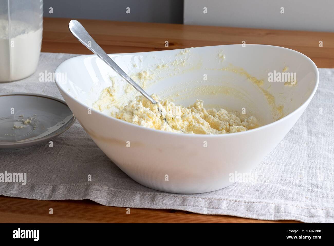Mixing cottage cheese dough in a white bowl with a fork on a linen tablecloth on a wooden table. Ingredients - cottage cheese, butter, egg and sugar. Stock Photo