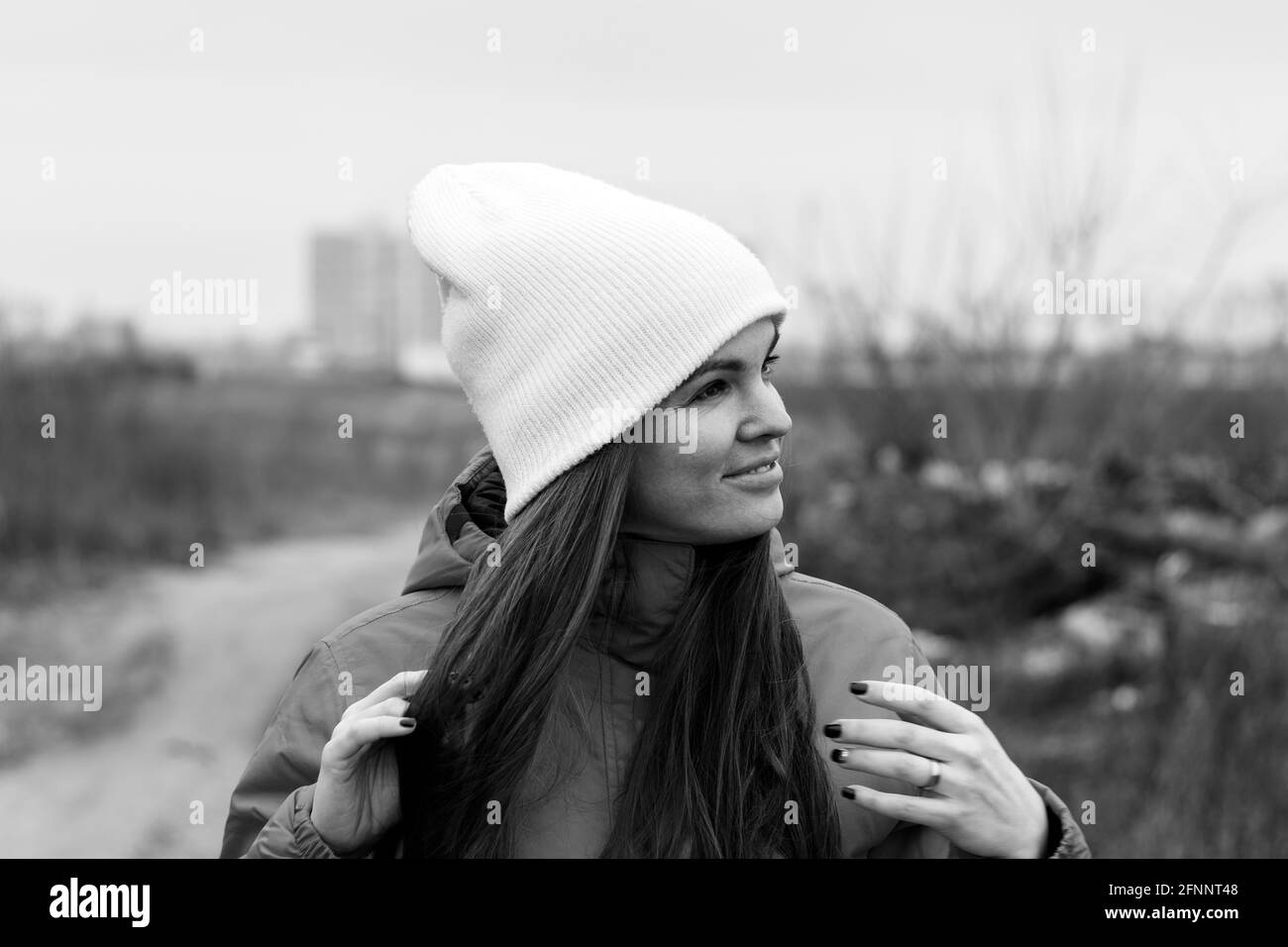 Smiling beautiful young woman with long hair in knitted hat looking away on the street on a cloudy day against a blurred background. Female without ma Stock Photo