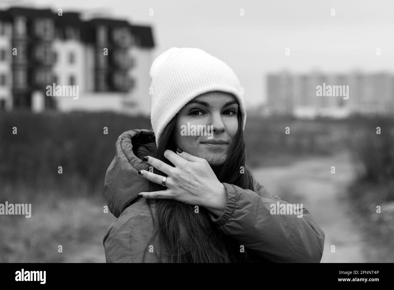 Smiling beautiful young woman with long hair in knitted hat on the street on a cloudy day against a blurred background. Female without makeup, natural Stock Photo