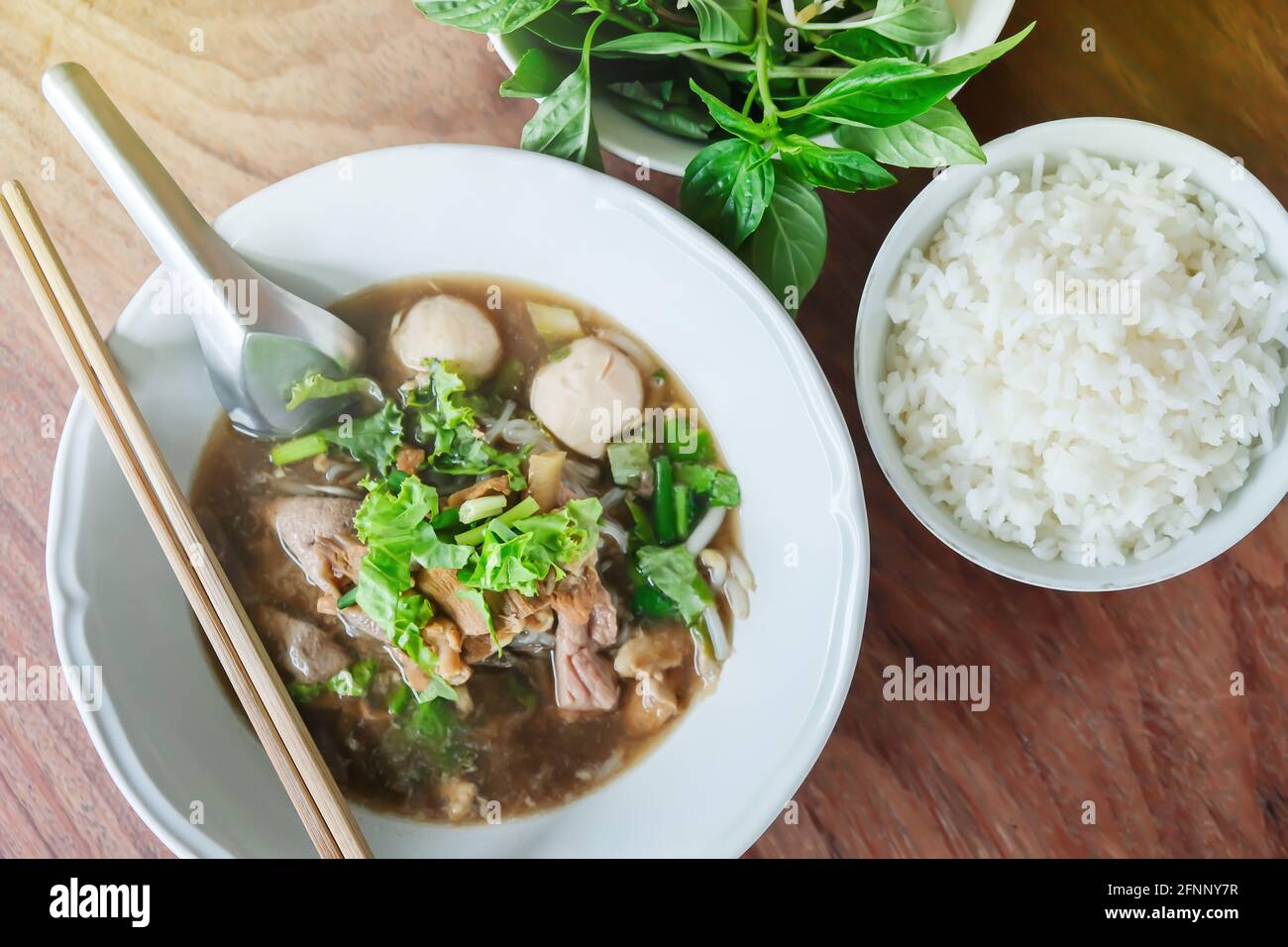 Braised beef noodle soup with pork ball on white bowl and steamed rice, basil leaves on wooden