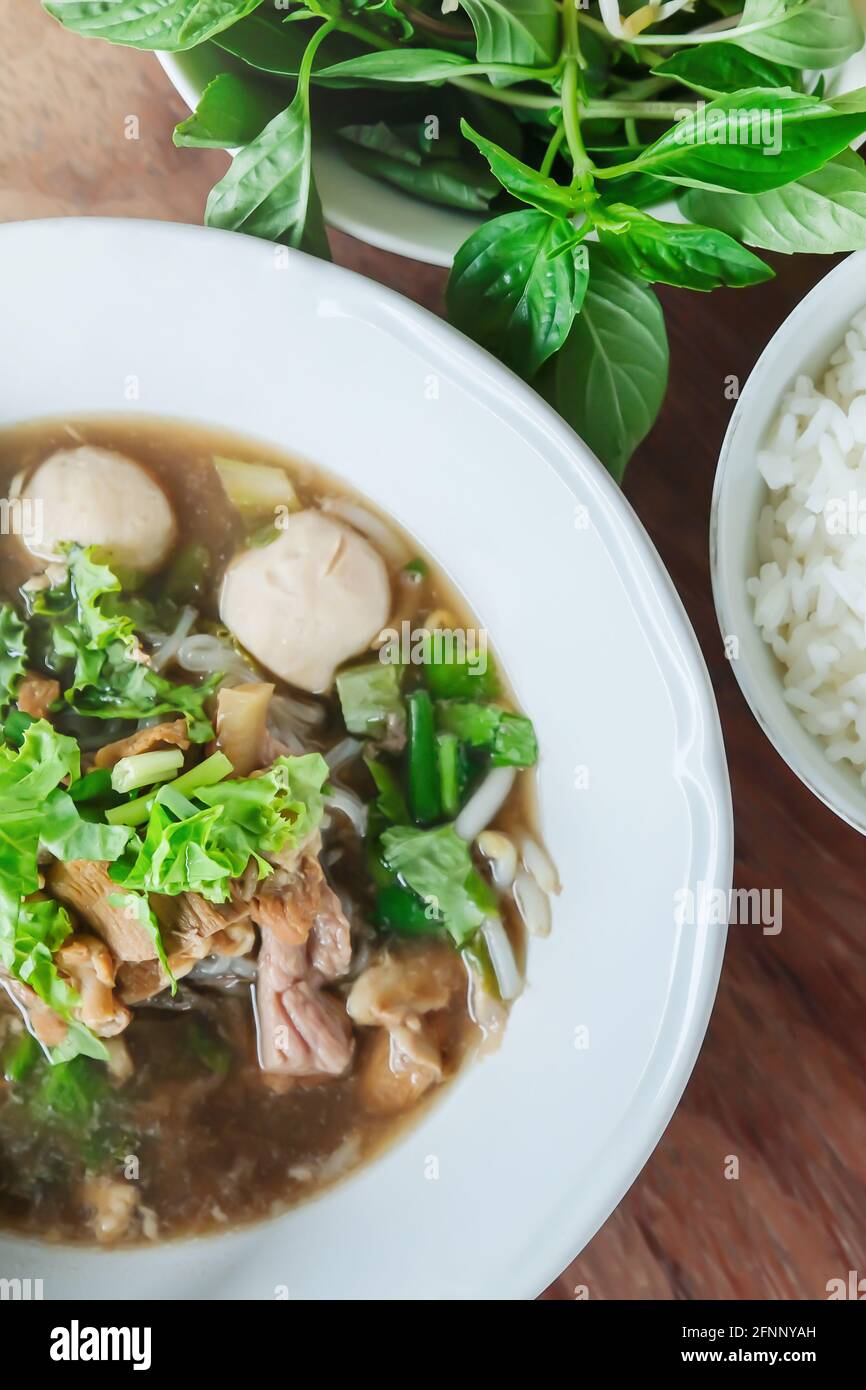Braised beef noodle soup with pork ball on white bowl and steamed rice, basil leaves on wooden