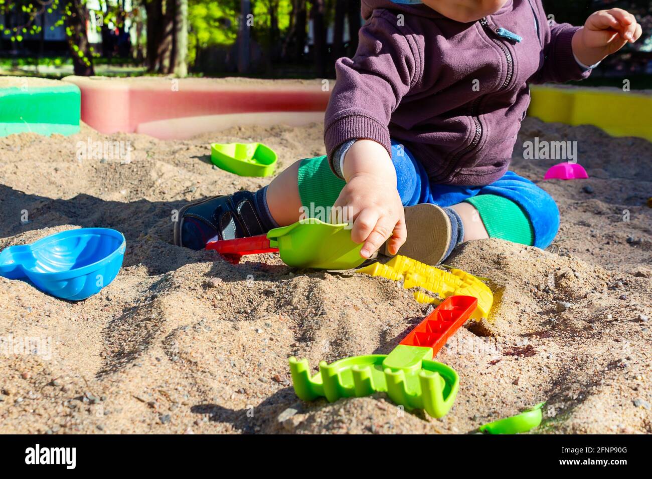 Unrecognizable little caucasian boy sitting in sandbox with toys on sunny summer day in big city. Child reaches for the bright plastic sand scoop.  Stock Photo