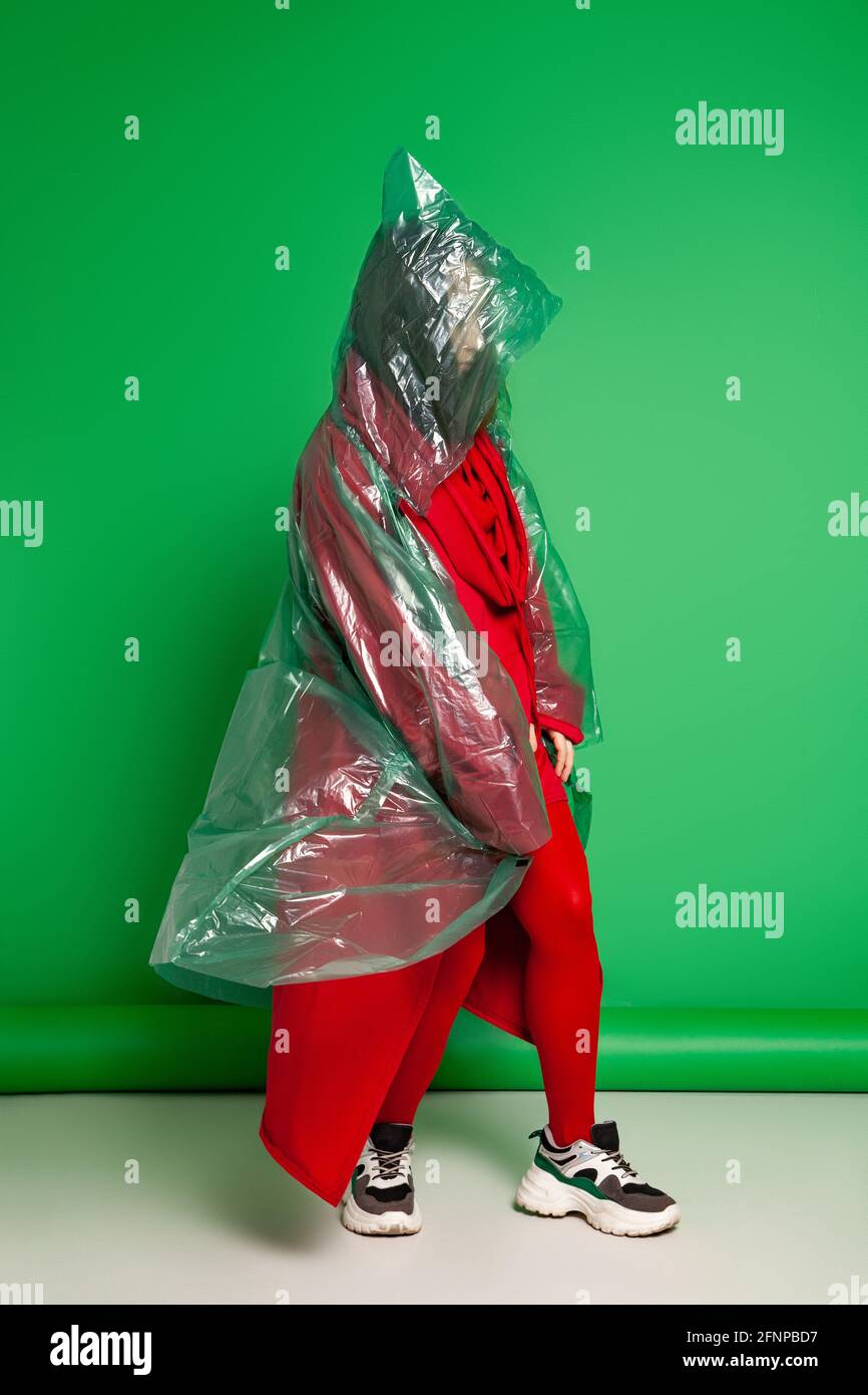 Calm female wearing transparent plastic raincoat and red clothes standing on green background in studio Stock Photo