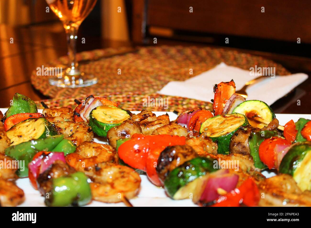 Close-up of grilled vegetable and shrimp skewers on a serving dish. Place setting with glass of wine out of focus in background. Stock Photo