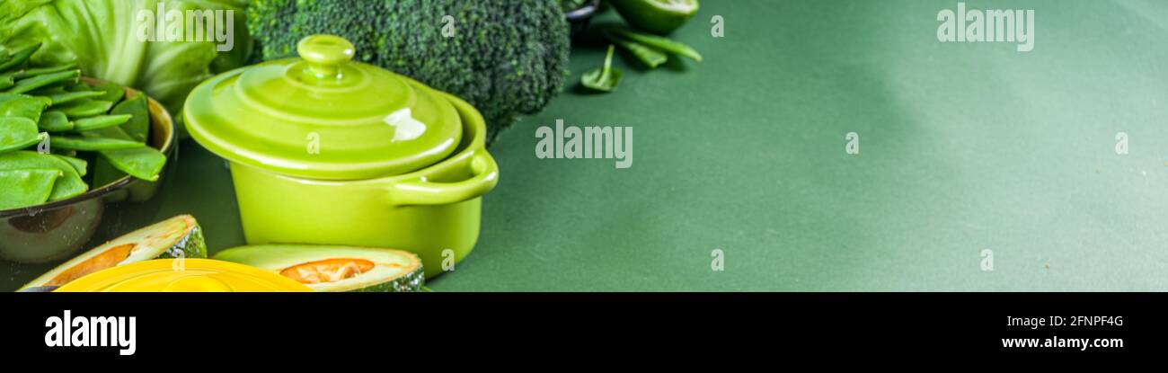 Various organic green spring vegetable ingredients with empty colorful portioned cooking pots on dark green kitchen table, top view. Diet healthy soup Stock Photo