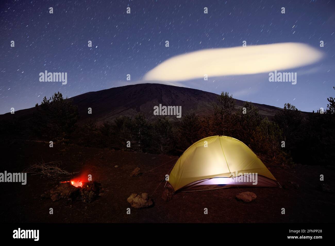 landscape in Sicily night with campfire and illuminated tent below Etna Volcano and star trails Stock Photo