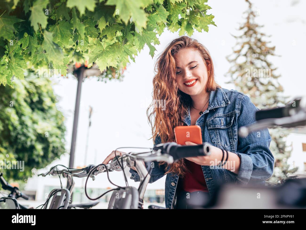 Portrait of smiling red curled long hair caucasian teen girl block out bike at Bicycle sharing point using the modern smartphone. Green urban transpor Stock Photo