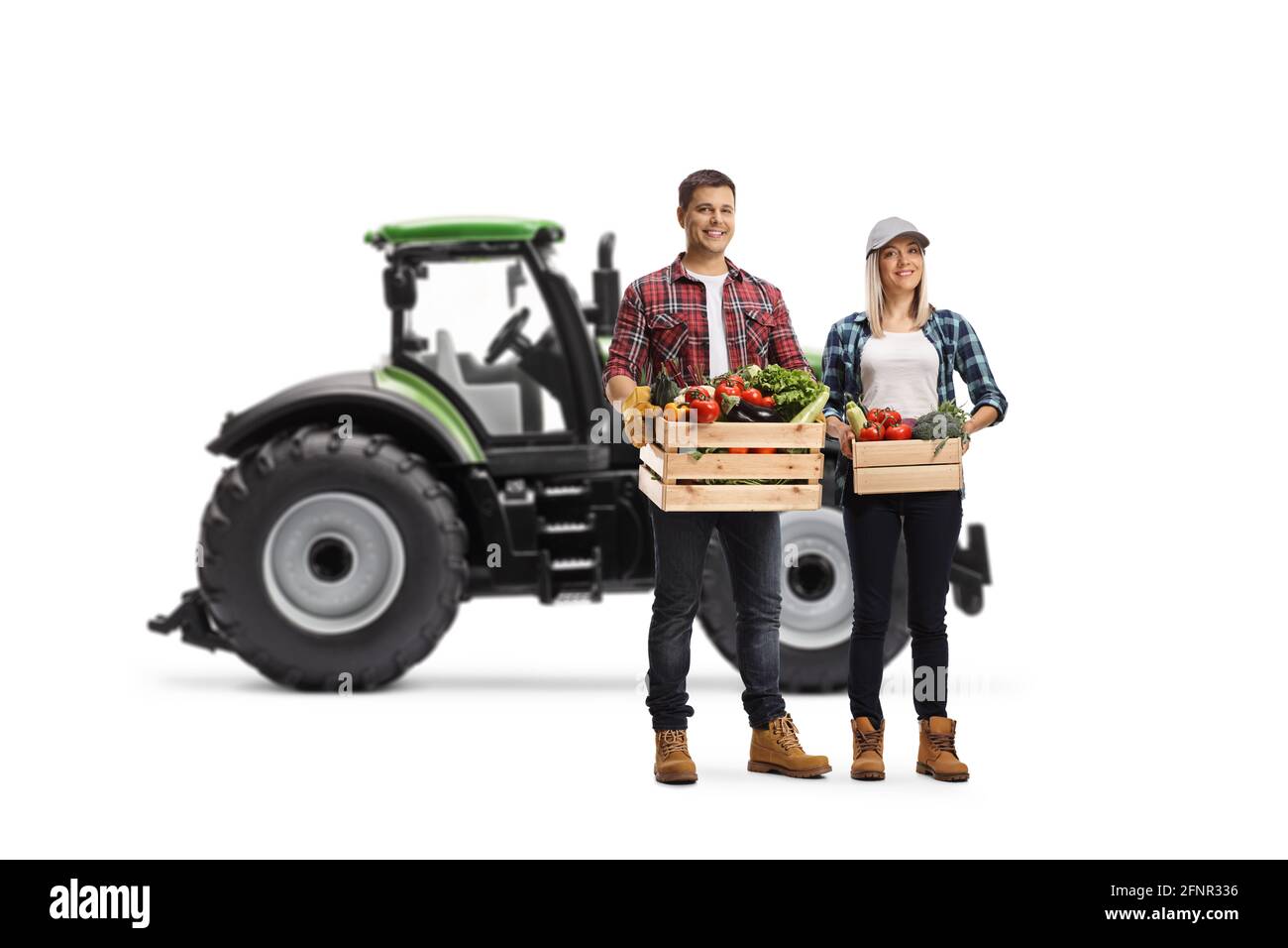 Young male and female farmers carrying crates with vegetables and posing in front of a tractor isolated on white background Stock Photo