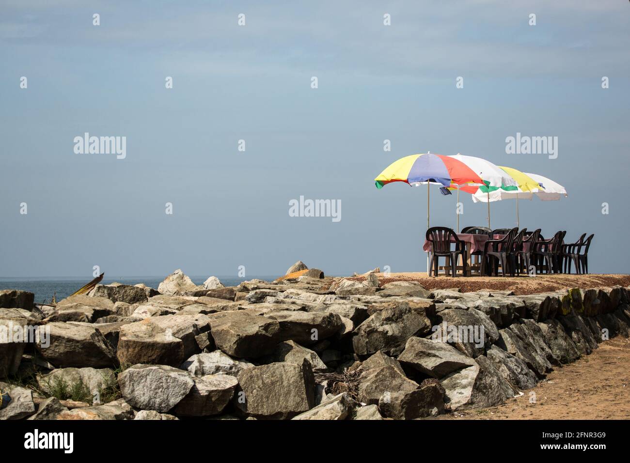 A group of chairs and tables under four colorful parasols stand on a heaped stone wall by the sea against a blue sky. Stock Photo