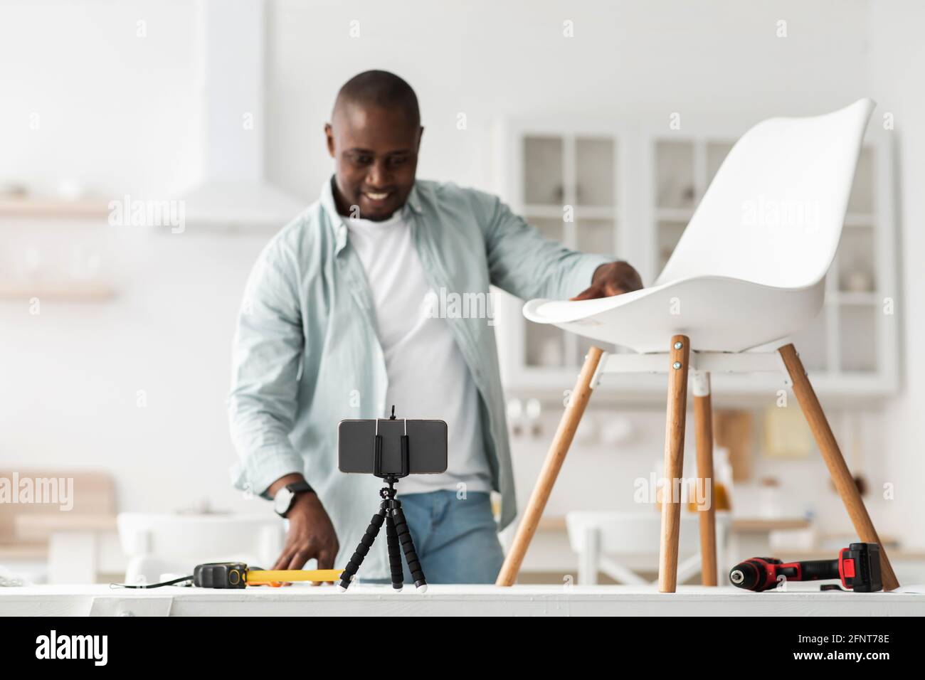 Modern blogger. Happy black man showing his ready-made chair and looking at smartphone camera, recording video Stock Photo