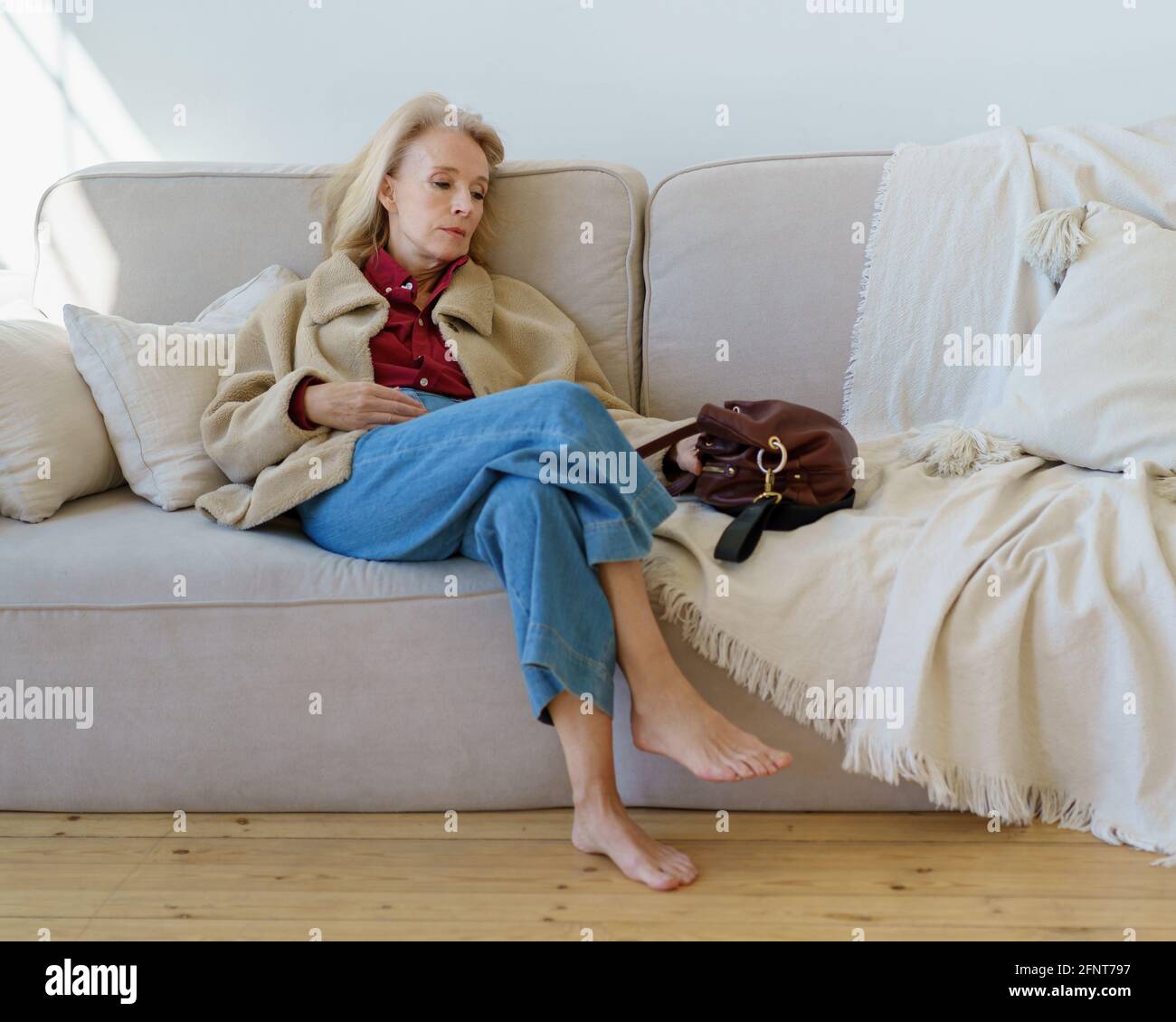 Tired mature 60s woman in casual wear resting after work on couch at home,  looking aside and thinking or pondering about something. Thoughtful senior  lady sitting on coach alone after hard working