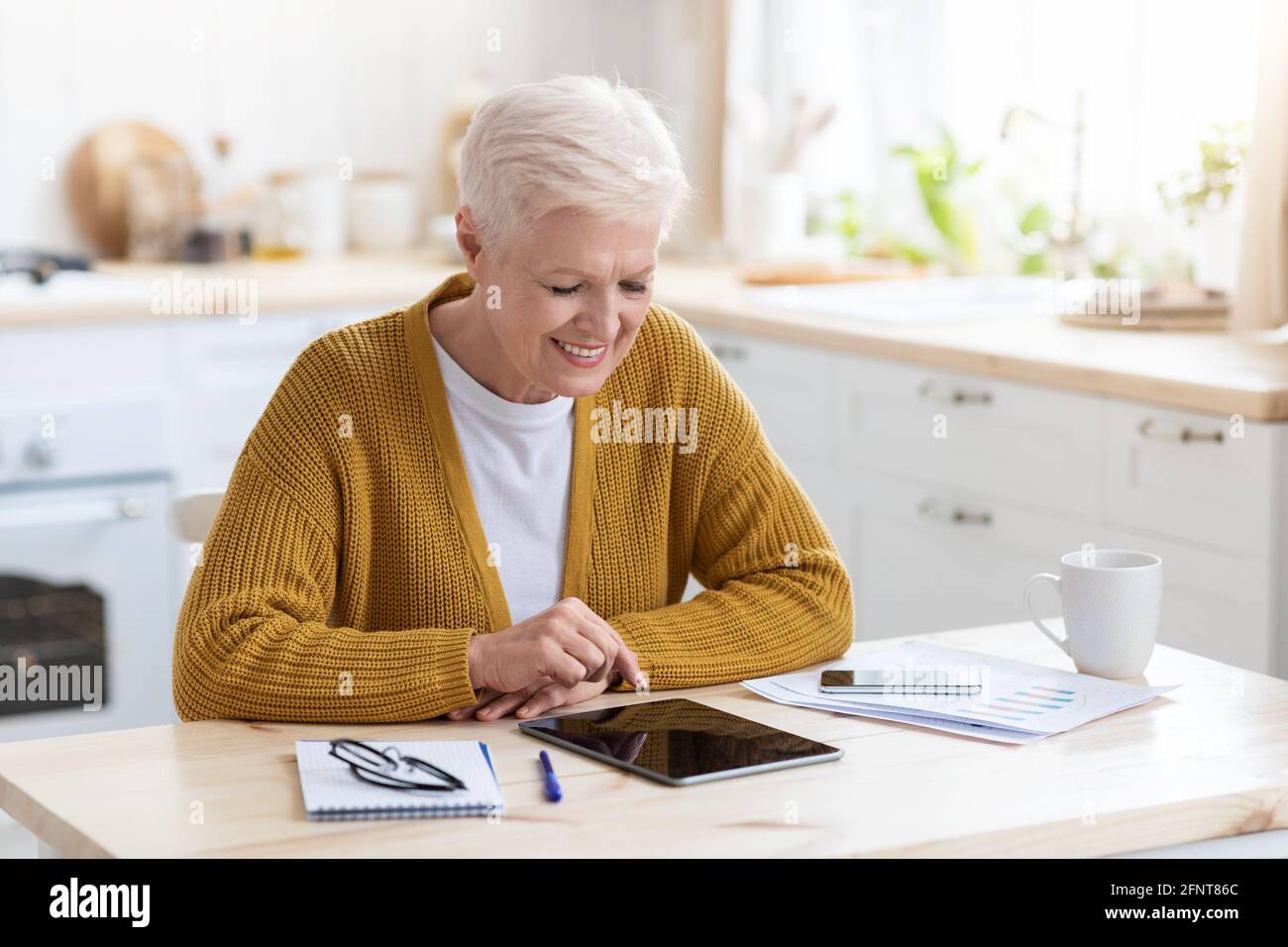 Joyful grey-haired senior woman using digital tablet in kitchen Stock Photo