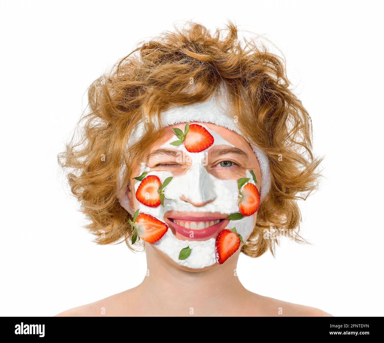 Red-haired curly young woman with a positive cheerful mood, smiling and winks, closed eye. With a cosmetic mask on the face of strawberries and cream. Stock Photo