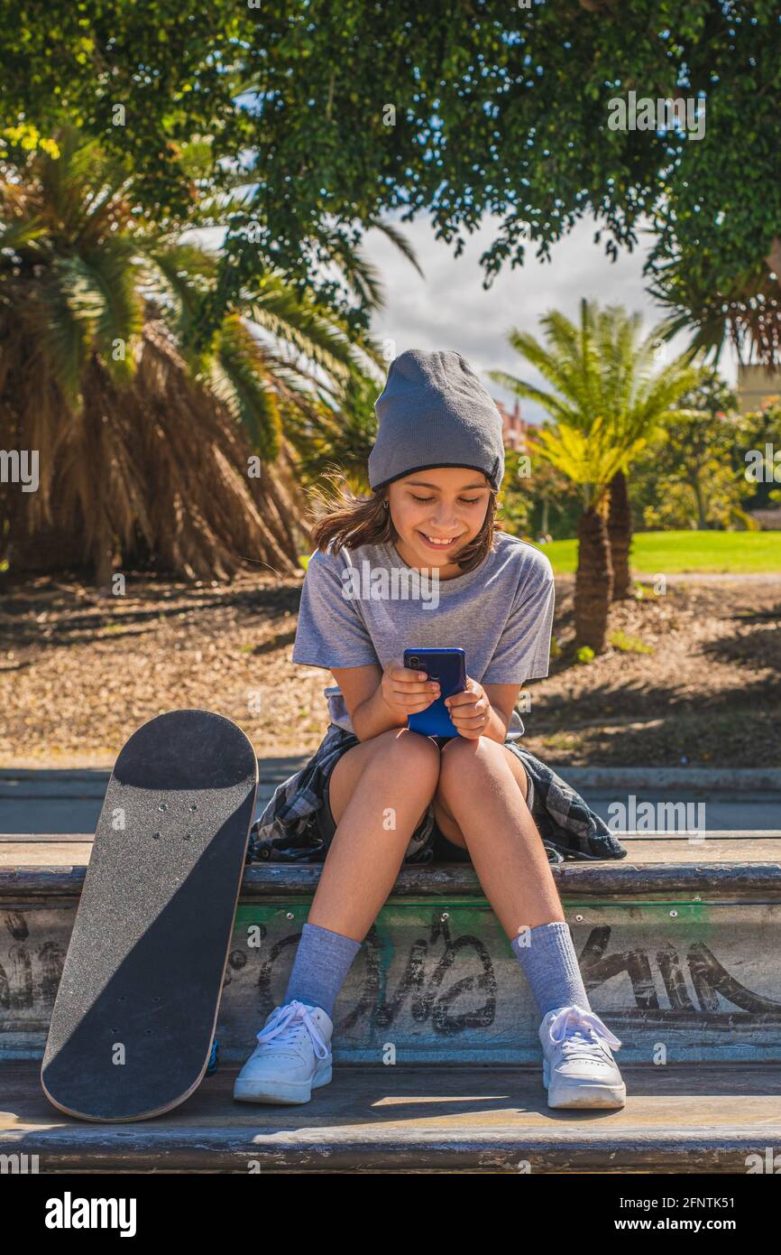 Pre-adolescent girl, Caucasian, sitting on an obstacle in a skate park, with her feet on her board, while chatting or surfing the internet with her mo Stock Photo