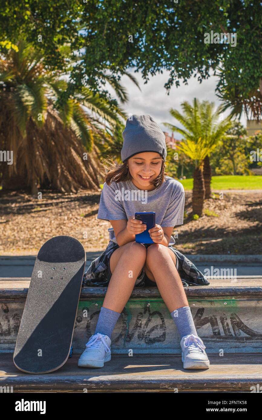 Pre-adolescent girl, Caucasian, sitting on an obstacle in a skate park, with her feet on her board, while chatting or surfing the internet with her mo Stock Photo