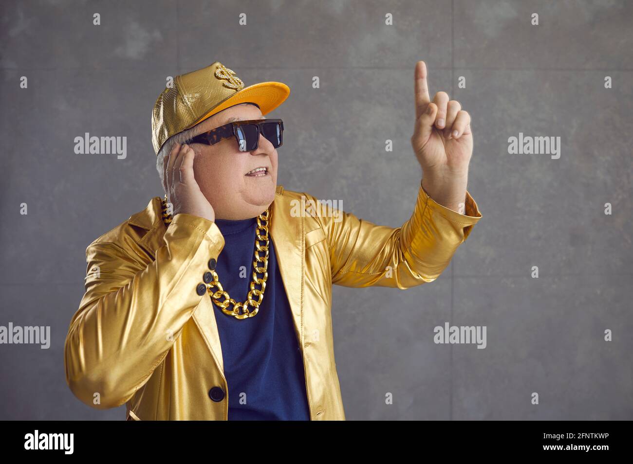 Funny senior DJ in golden jacket, baseball cap and chain necklace mixing music at disco party Stock Photo