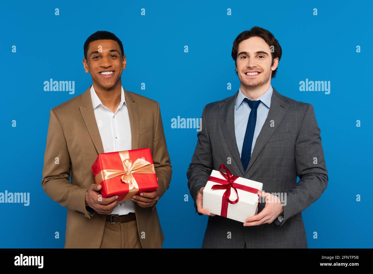 smiling young interracial men in fashionable suits holding gift boxes with red and golden ribbons isolated on blue Stock Photo