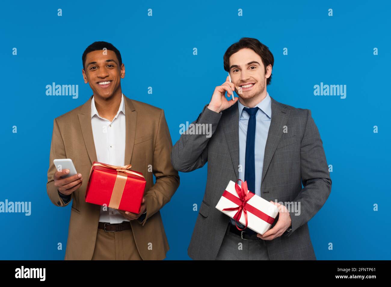smiling young interracial men in fashionable suits holding gift boxes with red and golden ribbons and using smartphones isolated on blue Stock Photo