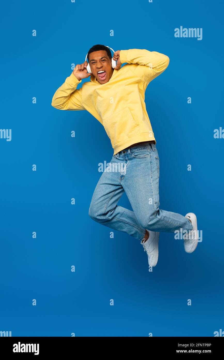 excited african american man jumping in headphones on blue background Stock Photo
