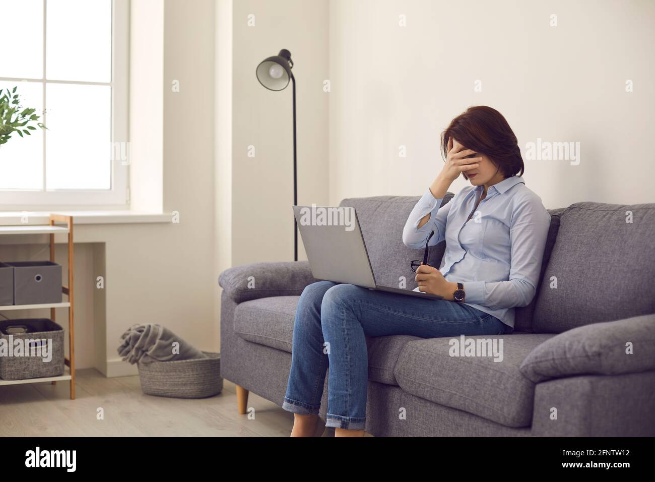 Woman sitting on sofa with laptop stops working and covers face feeling tired and unhappy Stock Photo
